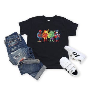 May include: A black t-shirt featuring a cartoon illustration of several superheroes, including Spider-Man, Iron Man, Hulk, and Captain America. The shirt is paired with blue denim jeans and white sneakers with black stripes.