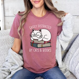 May include: A pink t-shirt with the text "Easily Distracted by Cats & Books" and a cartoon illustration of a white cat wearing glasses and sleeping on a stack of books.