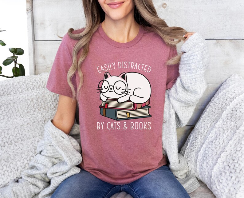 May include: A pink t-shirt with the text "Easily Distracted by Cats & Books" and a cartoon illustration of a white cat wearing glasses and sleeping on a stack of books.