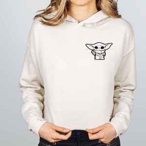 May include: A white cropped hoodie with a black and white illustration of a character from the Star Wars franchise. The character is wearing a hooded robe and has large eyes.