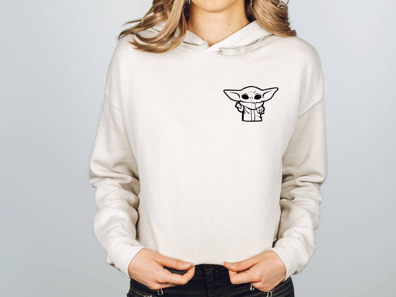 May include: A white cropped hoodie with a black and white illustration of a character from the Star Wars franchise. The character is wearing a hooded robe and has large eyes.