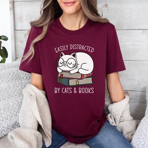 May include: A maroon t-shirt with the text "Easily Distracted by Cats & Books" and a cartoon illustration of a white cat wearing glasses and sleeping on top of a stack of books.