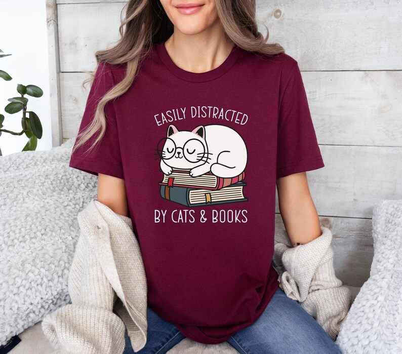 May include: A maroon t-shirt with the text "Easily Distracted by Cats & Books" and a cartoon illustration of a white cat wearing glasses and sleeping on top of a stack of books.