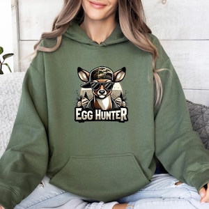 Puede incluir: Una sudadera con capucha de color verde oliva que presenta un estampado gráfico de un ciervo con una gorra de béisbol de camuflaje y gafas de sol negras. El texto EGG HUNTER está impreso en letras blancas en negrita debajo del ciervo. Mangas largas.
