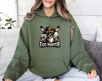 May include: An olive green hooded sweatshirt featuring a graphic print of a deer wearing a camouflage baseball cap and black sunglasses. The text EGG HUNTER is printed in bold white letters below the deer illustration. The hoodie has long sleeves.