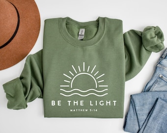 May include: An olive green crewneck sweatshirt featuring a white graphic of a rising sun over wavy lines. Below the graphic, the text reads BE THE LIGHT and MATTHEW 5:14 in a clean, sans-serif font. The sweatshirt has long sleeves and a ribbed collar.