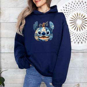 K&ouml;nnte beinhalten: Ein marineblauer Kapuzenpullover mit einem Stitch-inspirierten K&uuml;rbisdesign. Der K&uuml;rbis hat ein Patchworkmuster in Blau-, Braun- und Oranget&ouml;nen. Das Design befindet sich mittig auf der Vorderseite des Sweatshirts.