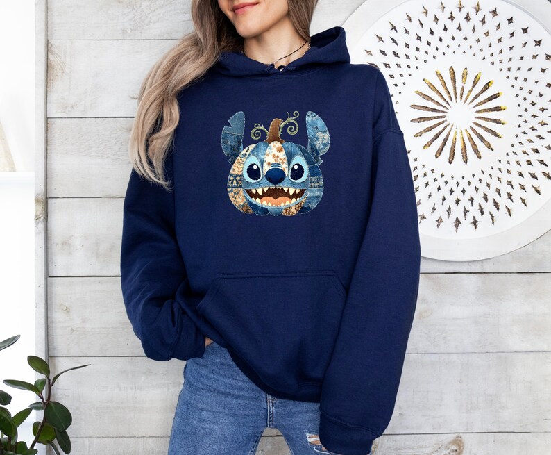 K&ouml;nnte beinhalten: Ein marineblauer Kapuzenpullover mit einem Stitch-inspirierten K&uuml;rbisdesign. Der K&uuml;rbis hat ein Patchworkmuster in Blau-, Braun- und Oranget&ouml;nen. Das Design befindet sich mittig auf der Vorderseite des Sweatshirts.