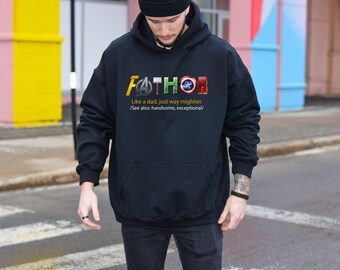 May include: A black hooded sweatshirt featuring the text FATHOR with superhero-themed icons replacing letters. Below the text, it reads: Like a dad, just way mightier /See also: handsome, exceptional/. The hoodie has long sleeves and a front pocket.
