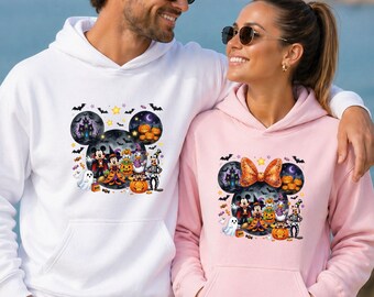 May include: A white hoodie and a light pink hoodie, each featuring a graphic print of cartoon characters in Halloween costumes, including a haunted house, pumpkins, bats, and a ghost, arranged within a large Mickey Mouse ear silhouette.