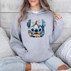 K&ouml;nnte beinhalten: Ein hellgrauer Kapuzenpullover mit einem von Stitch inspirierten K&uuml;rbisdesign. Der K&uuml;rbis hat blaue, braune und goldene Akzente mit einem l&auml;chelnden Gesicht. Der Pullover ist ein bequemer, l&auml;ssiger Stil.