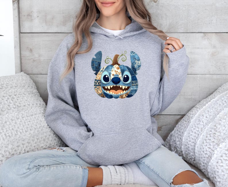 K&ouml;nnte beinhalten: Ein hellgrauer Kapuzenpullover mit einem von Stitch inspirierten K&uuml;rbisdesign. Der K&uuml;rbis hat blaue, braune und goldene Akzente mit einem l&auml;chelnden Gesicht. Der Pullover ist ein bequemer, l&auml;ssiger Stil.