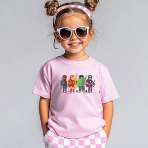 May include: A light pink t-shirt featuring a cartoon illustration of several superheroes, including Spider-Man, Iron Man, Hulk, and Captain America. The superheroes are holding pencils. The child is wearing sunglasses and a pink and white headband.