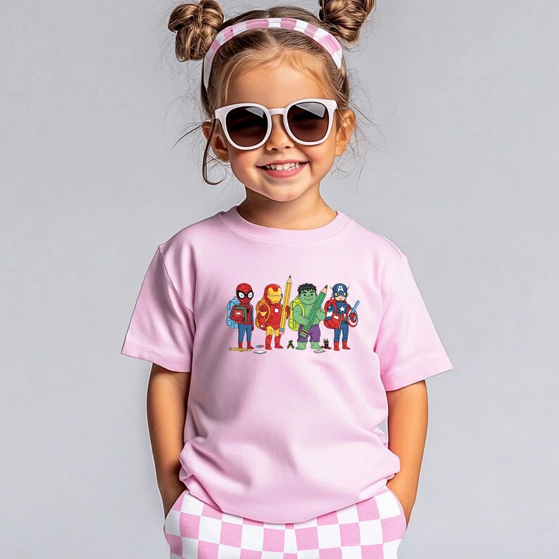 May include: A light pink t-shirt featuring a cartoon illustration of several superheroes, including Spider-Man, Iron Man, Hulk, and Captain America. The superheroes are holding pencils. The child is wearing sunglasses and a pink and white headband.