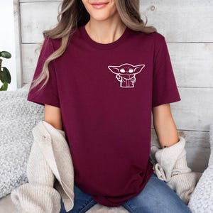 May include: A maroon t-shirt with a white outline of a character from the Star Wars franchise, "The Mandalorian".