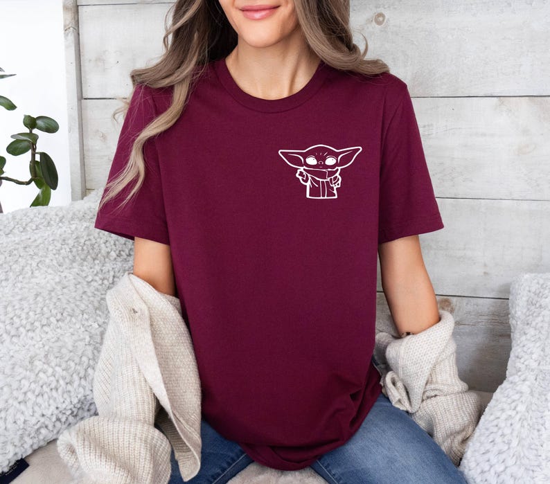 May include: A maroon t-shirt with a white outline of a character from the Star Wars franchise, "The Mandalorian".