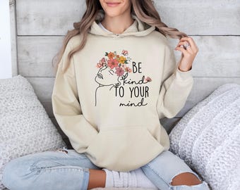 May include: A beige hooded sweatshirt featuring a black line art illustration of a profile face adorned with colorful flowers and butterflies. The text "BE kind TO YOUR mind" is printed in black lettering on the front of the garment.