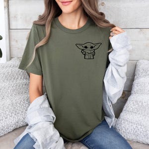 May include: A green t-shirt with a black graphic of a character from the Star Wars franchise. The character is known as "Baby Yoda" or "The Child".