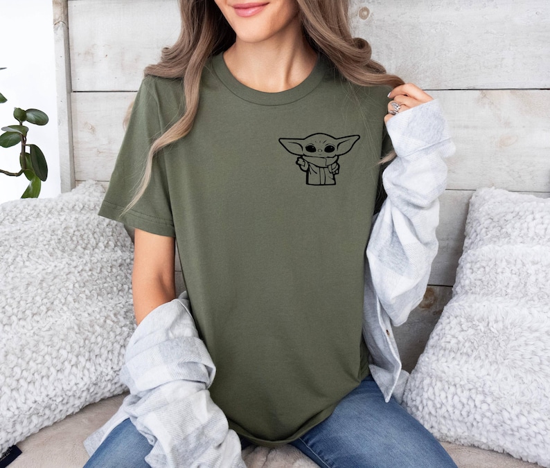 May include: A green t-shirt with a black graphic of a character from the Star Wars franchise. The character is known as "Baby Yoda" or "The Child".