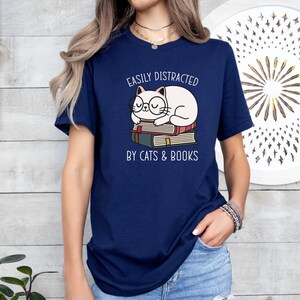 May include: A navy blue t-shirt with a white cat wearing glasses and sleeping on a stack of books. The text on the shirt reads "Easily Distracted by Cats & Books".