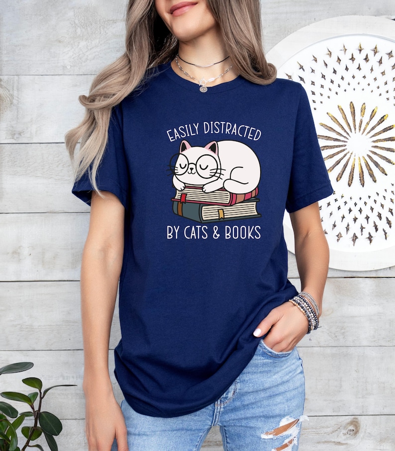 May include: A navy blue t-shirt with a white cat wearing glasses and sleeping on a stack of books. The text on the shirt reads "Easily Distracted by Cats & Books".