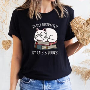 May include: Black t-shirt with a white cat sleeping on a stack of books. The text "Easily Distracted by Cats & Books" is printed on the shirt.