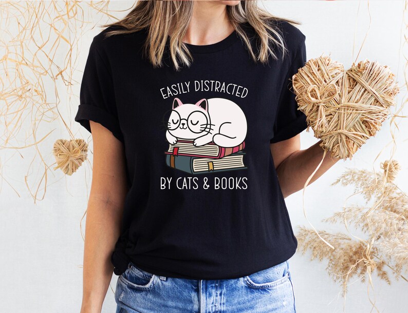 May include: Black t-shirt with a white cat sleeping on a stack of books. The text "Easily Distracted by Cats & Books" is printed on the shirt.
