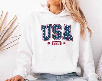 May include: A white hooded sweatshirt featuring a large graphic on the chest with the text USA in a blue block font filled with white stars and outlined in red. Below the text is a red rectangular badge containing the number 1776 with a star on each side.