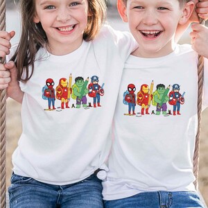 May include: Two children wearing white t-shirts with a colorful graphic of superheroes. The graphic features Spider-Man, Iron Man, Hulk, and Captain America, each holding school supplies. The children are smiling and appear to be on a swing.