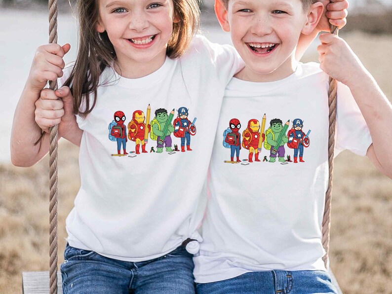 May include: Two children wearing white t-shirts with a colorful graphic of superheroes. The graphic features Spider-Man, Iron Man, Hulk, and Captain America, each holding school supplies. The children are smiling and appear to be on a swing.