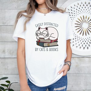 May include: White t-shirt with a graphic of a cat wearing glasses and sleeping on a stack of books. The text "Easily Distracted by Cats & Books" is printed below the cat.