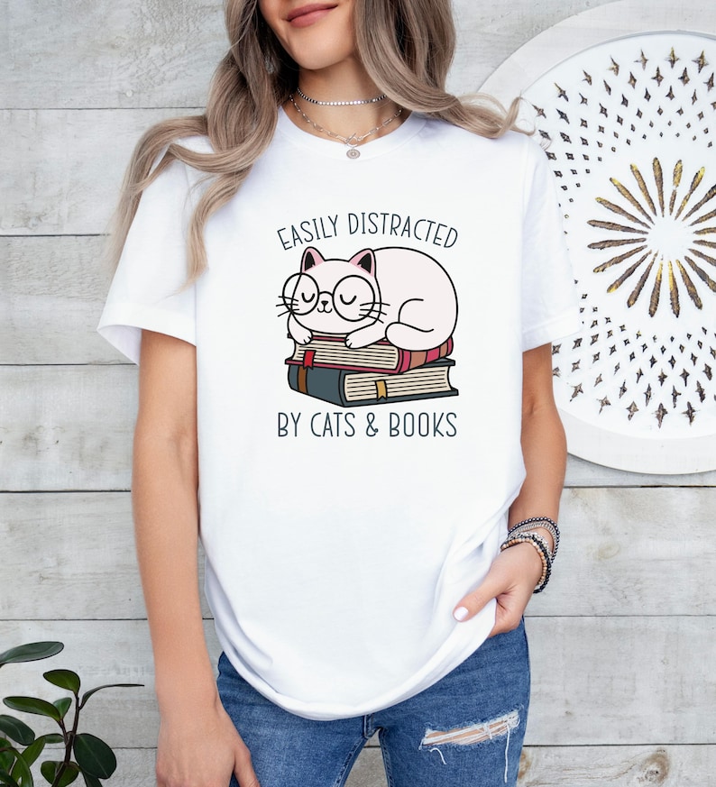 May include: White t-shirt with a graphic of a cat wearing glasses and sleeping on a stack of books. The text "Easily Distracted by Cats & Books" is printed below the cat.