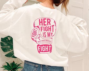 May include: A white long-sleeve sweatshirt featuring a large pink graphic on the back. The graphic depicts a boxing glove with the text "HER FIGHT IS MY FIGHT" and a small breast cancer awareness ribbon with the words "BREAST CANCER AWARENESS".