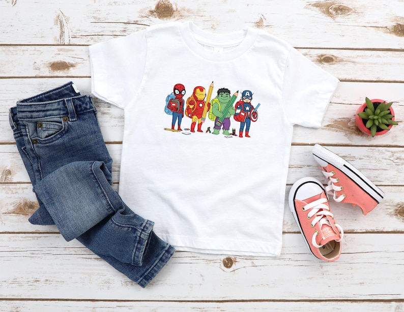 May include: White t-shirt featuring a cartoon illustration of superheroes, including Spider-Man, Iron Man, Hulk, Captain America, and others. The shirt is paired with blue jeans and coral-colored sneakers. A small potted plant is also visible.