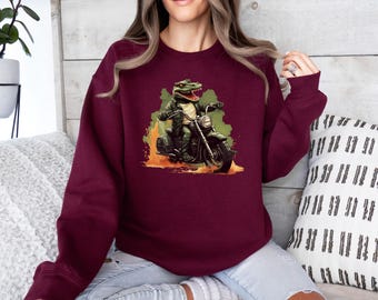 May include: A maroon long-sleeve crewneck sweatshirt featuring a graphic print of a green alligator wearing a black leather jacket and riding a motorcycle. The design includes orange and green abstract shapes behind the alligator and motorcycle.