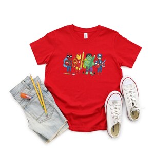 May include: A red t-shirt featuring a cartoon illustration of superheroes, including Spider-Man, Iron Man, Hulk, and Captain America, each holding school supplies. The shirt is paired with denim shorts, pencils, glue stick, and white sneakers.