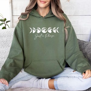 May include: An olive green hooded sweatshirt featuring a white graphic of the lunar cycle phases across the chest. Below the moon phases, the text "Just a Phase" is printed in a white cursive script. The garment includes a front kangaroo pocket.