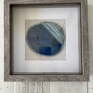 May include: A framed agate slice with a blue and white pattern. The agate is displayed in a gray wood frame with a white mat.