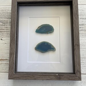 May include: A framed artwork featuring two agate slices with a teal and green color scheme. The agate slices are displayed on a white background within a dark gray wood frame.