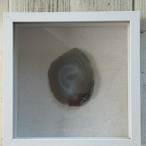 May include: A square white shadow box frame containing a single, polished, translucent agate slice. The stone features concentric bands of light gray, beige, and white, set against a neutral, textured, off-white fabric backing.
