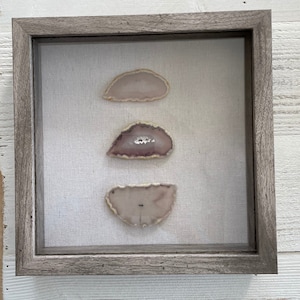 May include: Three agate slices in a wooden frame. The agate slices are in shades of pink and white. The frame is a light gray color.