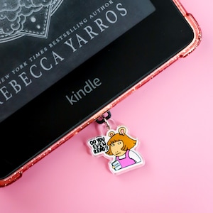 May include: A Kindle e-reader with a pink glitter case displaying the book "Iron Flame" by Rebecca Yarros. A charm featuring a cartoon character with the text "DO YOU EVEN READ?!" hangs from the device, set against a pink background.