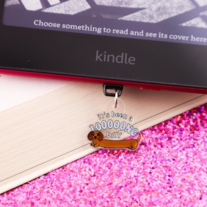 May include: A Kindle e-reader with a red border rests on a book, with a dachshund-shaped bookmark that reads "It's been a 100000NG DAY." The background is a sparkling pink surface.