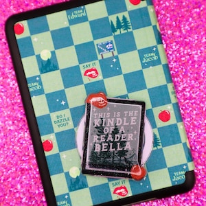 May include: A black and green checkered phone case with a black and white sticker that says "This is the Kindle of a Reader, Bella". The sticker has a red lip design on the top and bottom.