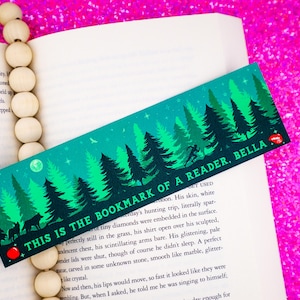 May include: A teal blue bookmark with a forest scene and the text "This is the bookmark of a reader. Bella."  The bookmark is laying on a book with a wooden bead necklace.