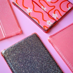 May include: Four phone cases with glitter on a pink background. One case has a pink and orange abstract design with white flowers. Another case is black with a glittery finish. The other two cases are pink with a glittery finish.