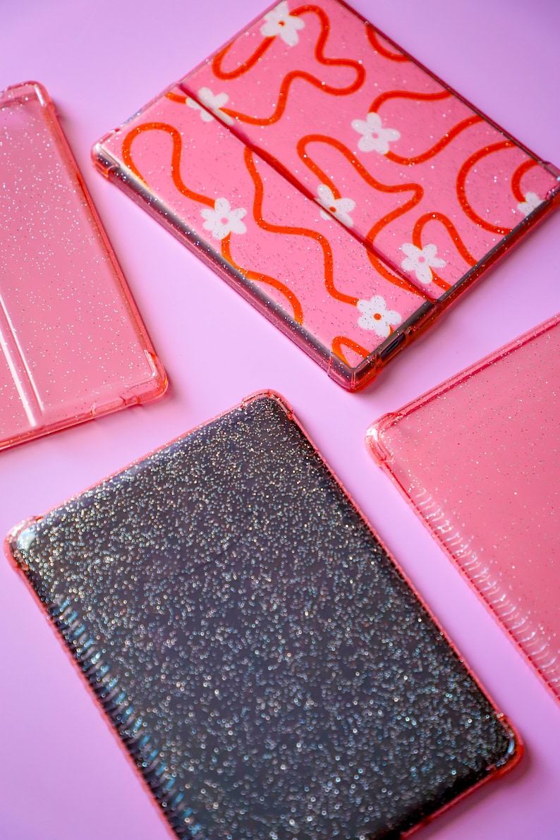 May include: Four phone cases with glitter on a pink background. One case has a pink and orange abstract design with white flowers. Another case is black with a glittery finish. The other two cases are pink with a glittery finish.