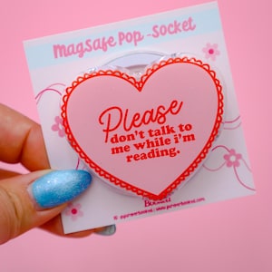 May include: A pink heart-shaped MagSafe Pop-Socket with a red scalloped border. The text "Please don't talk to me while I'm reading" is written in red. The background is pink.