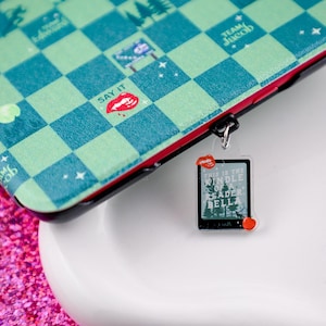 May include: A green and blue checkered phone case with a red lip and the words "Say It" printed on it. A clear charm with the words "This is the middle of a reader Bella" is attached to the case.