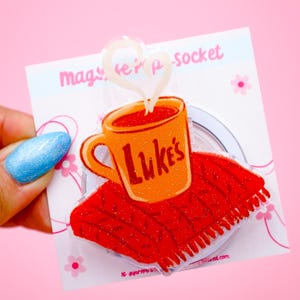 May include: A phone grip featuring a coffee cup design with the text "Luke's" on a red knitted coaster. The grip has a heart-shaped steam design and is attached to a white backing with text and pink flower accents.